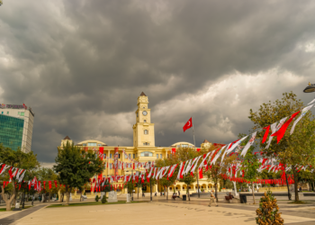 Şehir meydanında Türk bayrakları asılı tarihi belediye binası ve bulutlu gökyüzü.