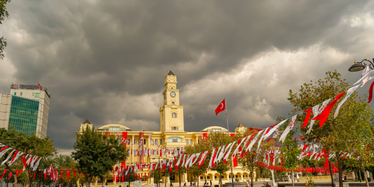 Şehir meydanında Türk bayrakları asılı tarihi belediye binası ve bulutlu gökyüzü.