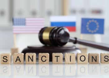 Ahşap harflerle yazılmış sanctions yazısı arkasında küçük bayraklar ve tokmak duruyor.