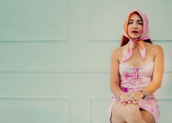 Açık mavi duvar önünde oturan, pembe desenli elbiseli ve eşarp takmış kadın