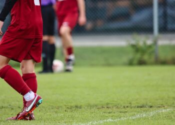 Kırmızı formalı futbolcunun çimenler üzerindeki kramponu ve bacağı.