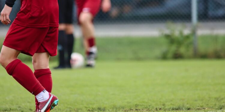 Kırmızı formalı futbolcunun çimenler üzerindeki kramponu ve bacağı.