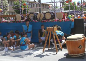 Bir festival sahnesi önünde yöresel kıyafetli Japon davulcular oturuşmaktadır.