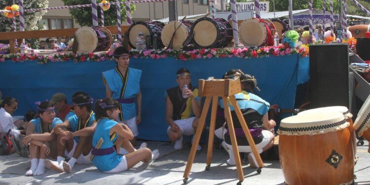 Bir festival sahnesi önünde yöresel kıyafetli Japon davulcular oturuşmaktadır.