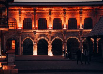 Gece ışıklandırılmış tarihi taş binanın avlusu ve kemerli sütunları.