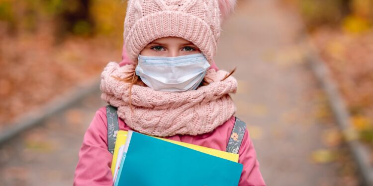 Pembe mont ve şapka giymiş, maske takıp kitaplar tutan kız çocuğu.