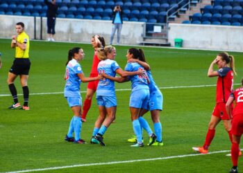 Mavi formalar giyen kadın futbolcular sahada sevinçle birbirine sarılıyor.