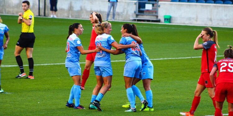 Mavi formalar giyen kadın futbolcular sahada sevinçle birbirine sarılıyor.
