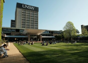 Geniş çim alanda oturan öğrencilerin arkasında UNSW yazılı büyük üniversite binası.