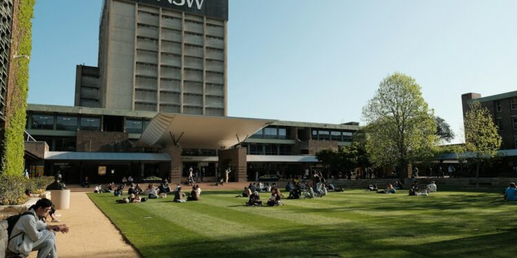 Geniş çim alanda oturan öğrencilerin arkasında UNSW yazılı büyük üniversite binası.