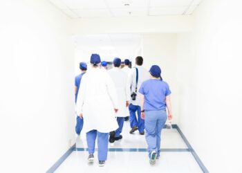 Hastane koridorunda yürüyen mavi ve beyaz üniformalı sağlık personeli ekibi.