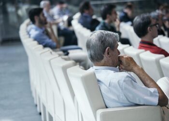 Konferans salonunda koltuklarda oturup düşünen katılımcıların yandan görünümü.