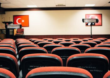 Kırmızı koltuklu boş bir konferans salonu ve arka duvarda Türk bayrakları asılı.